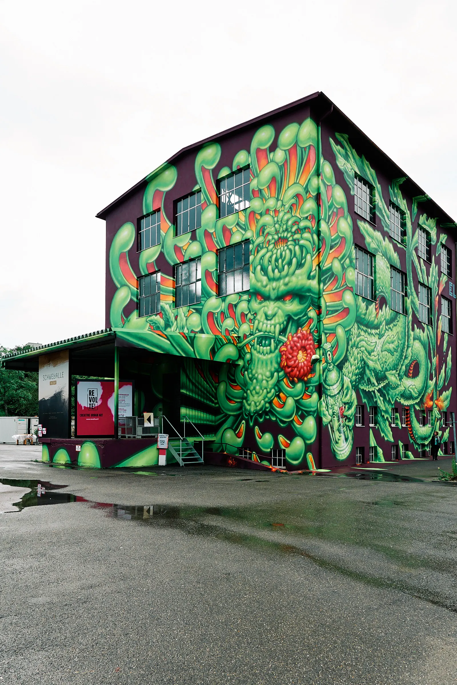 Graffito - Giftgrünes mit orangefarbenen Elementen auf dunklem, lilafarbenem Grund, der Phantasiewelt entspringendes Wesen, dass wie eine Kombination dreier Lebensformen - Drache, Krake, Blütenkelch - wirkt und eine Spraydose in seinen „Tentakeln“ hält. Das Graffito geht vom Boden bis unter das Dach am Eingang der circa 20 Meter hohen Schwelhalle Balingen und über Eck auf zwei Fassadenseiten.